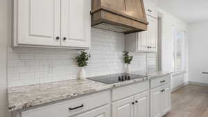 Kitchen with white cabinetry, custom range hood, black electric stovetop, backsplash, and light wood-type flooring