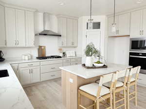 Kitchen featuring light wood finished floors, appliances with stainless steel finishes, hanging light fixtures, light stone countertops, and wall chimney range hood