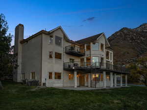 Back house at dusk featuring a lawn, a balcony, central AC unit, and a patio area