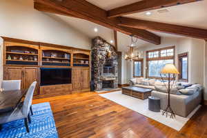 Living room with a chandelier, wood-type flooring, vaulted ceiling with beams, and a stone fireplace