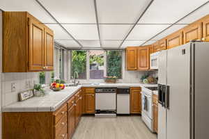 Kitchen with tile countertops, white appliances, sink, light wood-type flooring, and tasteful backsplash