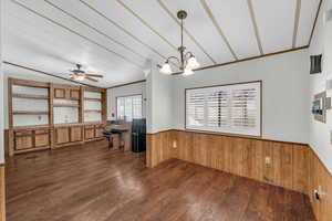 Interior space featuring dark hardwood / wood-style flooring, pendant lighting, wooden walls, ceiling fan with notable chandelier, and ornamental molding