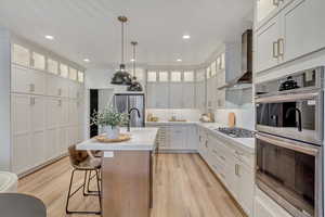 Kitchen with a kitchen island with sink, white cabinets, wall chimney range hood, light wood-type flooring, and stainless steel appliances