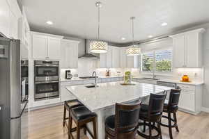 Kitchen with stainless steel appliances, sink, wall chimney range hood, a center island with sink, and white cabinetry