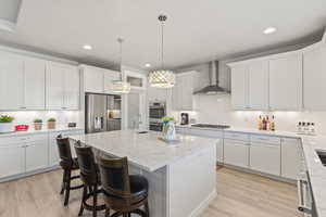 Kitchen featuring wall chimney range hood, an island with sink, light hardwood / wood-style floors, white cabinetry, and stainless steel appliances