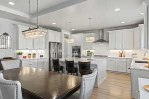 Kitchen featuring white cabinets, an island with sink, decorative light fixtures, and wall chimney range hood