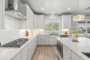 Kitchen featuring appliances with stainless steel finishes, light wood-type flooring, wall chimney exhaust hood, decorative light fixtures, and white cabinetry