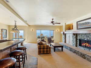 Living room featuring a stone fireplace, ceiling fan, bar, beam ceiling, and carpet floors