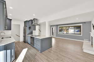 Kitchen featuring stainless steel appliances, wall chimney range hood, light stone countertops, sink, and light hardwood / wood-style flooring