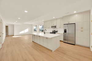 Kitchen featuring appliances with stainless steel finishes, light wood-type flooring, sink, white cabinetry, and an island with sink