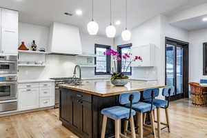 Kitchen with custom range hood, stainless steel double oven, a center island with sink, white cabinets, and light hardwood / wood-style floors