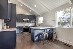 Kitchen with appliances with stainless steel finishes, a kitchen breakfast bar, blue cabinetry, vaulted ceiling with beams, and dark hardwood / wood-style floors