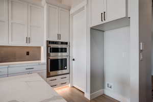 Kitchen with light stone countertops, light wood-type flooring, backsplash, double oven, and white cabinets