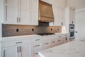 Kitchen with light stone countertops, backsplash, custom exhaust hood, stainless steel appliances, and white cabinets