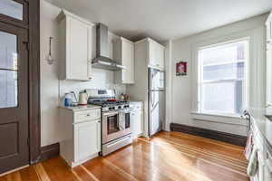 Kitchen with white cabinetry, wall chimney exhaust hood, light hardwood / wood-style floors, and appliances with stainless steel finishes