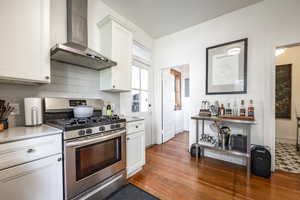 Kitchen with wall chimney exhaust hood, stainless steel gas range oven, white cabinetry, and dark hardwood / wood-style floors