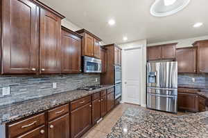 Kitchen with dark stone counters, appliances with stainless steel finishes, tasteful backsplash, dark brown cabinets, and light tile patterned flooring