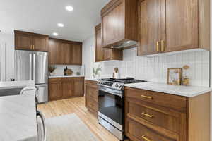 Kitchen with decorative backsplash, light stone counters, wall chimney exhaust hood, stainless steel appliances, and light hardwood / wood-style floors