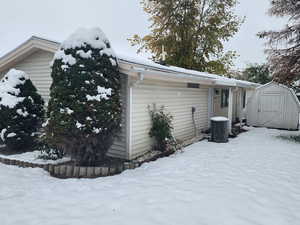 View of snow covered exterior with central AC unit and a storage shed