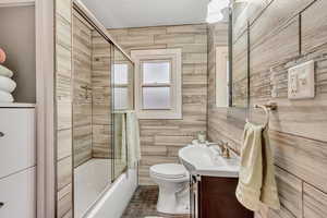 Full bathroom with enclosed tub / shower combo, vanity, toilet, and wooden walls