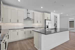 Kitchen with appliances with stainless steel finishes, an island with sink, white cabinetry, and wall chimney range hood (work in progress),