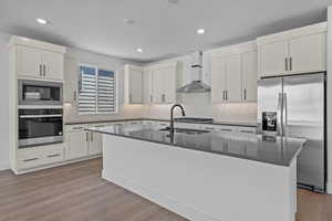 Kitchen featuring appliances with stainless steel finishes, sink, wall chimney range hood, a center island with sink, and light hardwood / wood-style flooring