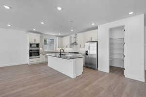 Kitchen with stainless steel appliances, sink, wall chimney range hood, a center island with sink, and light hardwood / wood-style flooring