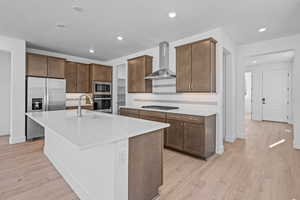 Kitchen with sink, stainless steel appliances, wall chimney range hood, an island with sink, and light hardwood / wood-style floors