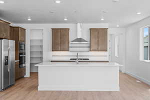 Kitchen with a center island with sink, light hardwood / wood-style flooring, wall chimney range hood, and appliances with stainless steel finishes