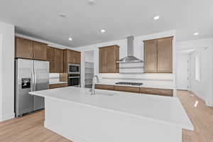 Kitchen with wall chimney exhaust hood, stainless steel appliances, sink, light hardwood / wood-style floors, and an island with sink