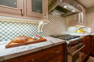 Kitchen with backsplash, crown molding, wall chimney exhaust hood, high end stove, and light stone counters