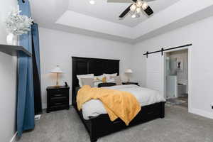 Carpeted bedroom featuring a raised ceiling, a barn door, ceiling fan, and ensuite bath