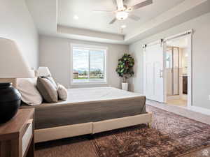 Bedroom featuring a tray ceiling, a barn door, ceiling fan, and connected bathroom