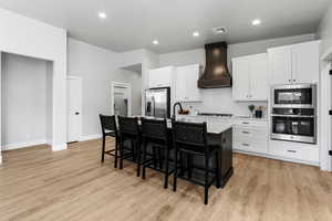 Kitchen featuring stainless steel appliances, custom exhaust hood, a breakfast bar area, a kitchen island with sink, and light wood-style flooring