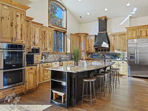 Kitchen featuring light stone counters, custom exhaust hood, a kitchen island with sink, built in appliances, and high vaulted ceiling