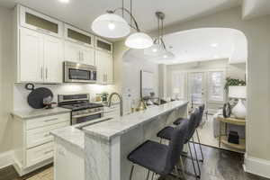 Kitchen with dark wood-type flooring, light stone countertops, an island with sink, decorative light fixtures, and stainless steel appliances