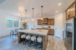 Kitchen featuring stainless steel appliances, decorative light fixtures, a kitchen island with sink, light wood-type flooring, and a breakfast bar