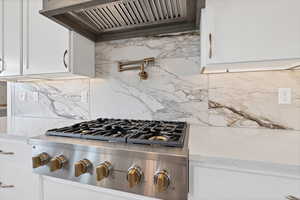 Kitchen view of stainless steel gas cooktop, custom range hood, pot filler, decorative backsplash, and white cabinets