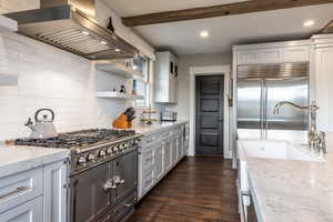 Kitchen with light stone countertops, wall chimney exhaust hood, dark wood-type flooring, beam ceiling, and built in appliances