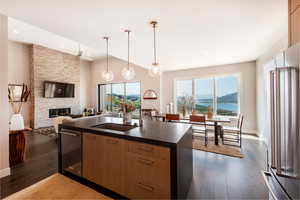 Kitchen with dark wood-type flooring, a stone fireplace, high end refrigerator, decorative light fixtures, and a kitchen island with sink