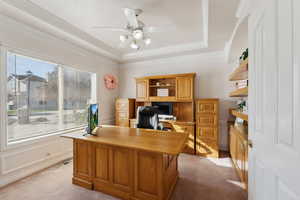 Carpeted home office featuring ceiling fan, a raised ceiling, built in desk, and crown molding