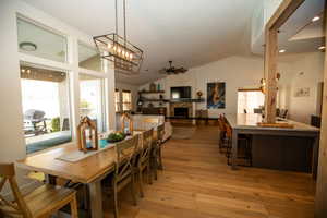 Dining room with a stone fireplace, ceiling fan with notable chandelier, vaulted ceiling, and light wood-type flooring