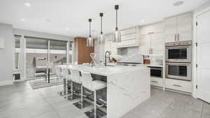 Kitchen featuring backsplash, hanging light fixtures, an island with sink, light stone counters, and stainless steel appliances