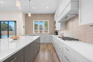 Kitchen featuring light hardwood / wood-style floors, stainless steel gas cooktop, white cabinetry, custom range hood, and pendant lighting