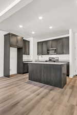 Kitchen featuring light hardwood / wood-style floors, dark brown cabinetry, an island with sink, and appliances with stainless steel finishes