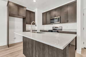 Kitchen with light wood-type flooring, light stone counters, stainless steel appliances, a kitchen island with sink, and sink