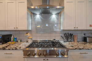 Kitchen with tasteful backsplash, light stone counters, stainless steel gas cooktop, wall chimney range hood, and white cabinetry