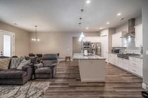 Kitchen featuring wall chimney exhaust hood, stainless steel appliances, pendant lighting, a kitchen island with sink, and white cabinets