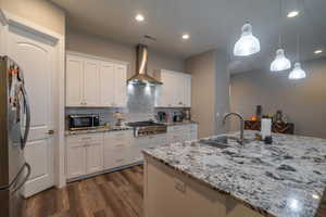 Kitchen with white cabinets, wall chimney range hood, hanging light fixtures, sink, and appliances with stainless steel finishes