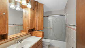 Full bathroom featuring enclosed tub / shower combo, a textured ceiling, toilet, vanity, and tile walls